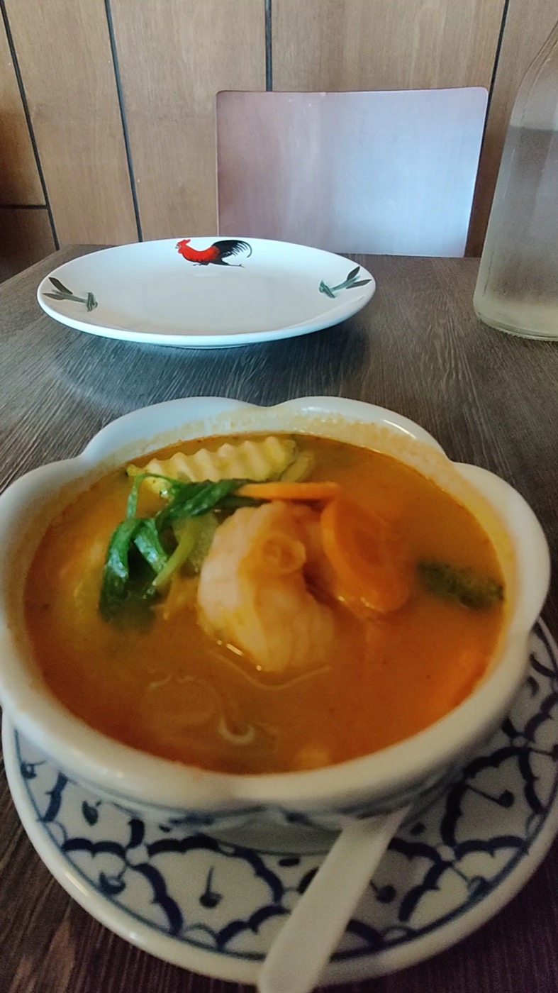 A blue-and-white bowl of orange broth with prawns, baby corn and fresh herbs on a wooden table