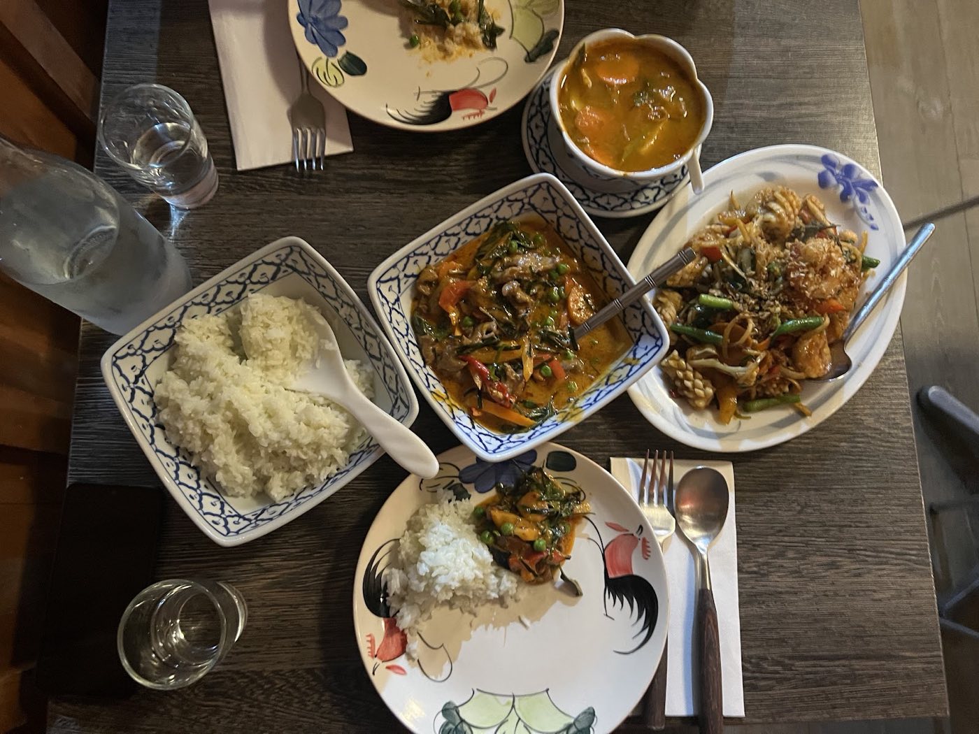 Overhead view of a dark table with a bowl of rice, a curry, a stir-fried noodle dish and a small bowl of soup on patterned plates