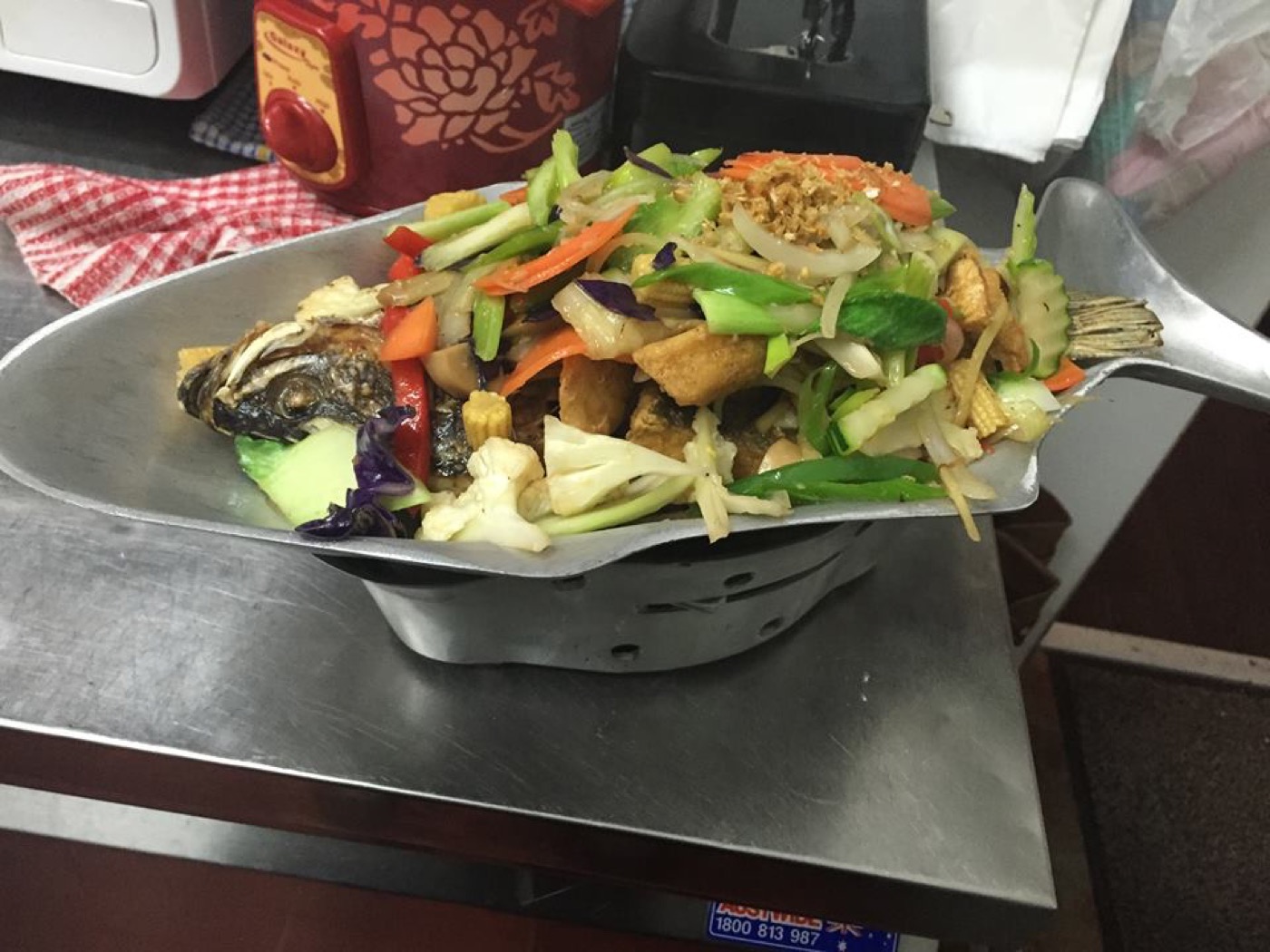 A whole fish on a metal platter over a small burner, covered with a pile of stir-fried vegetables